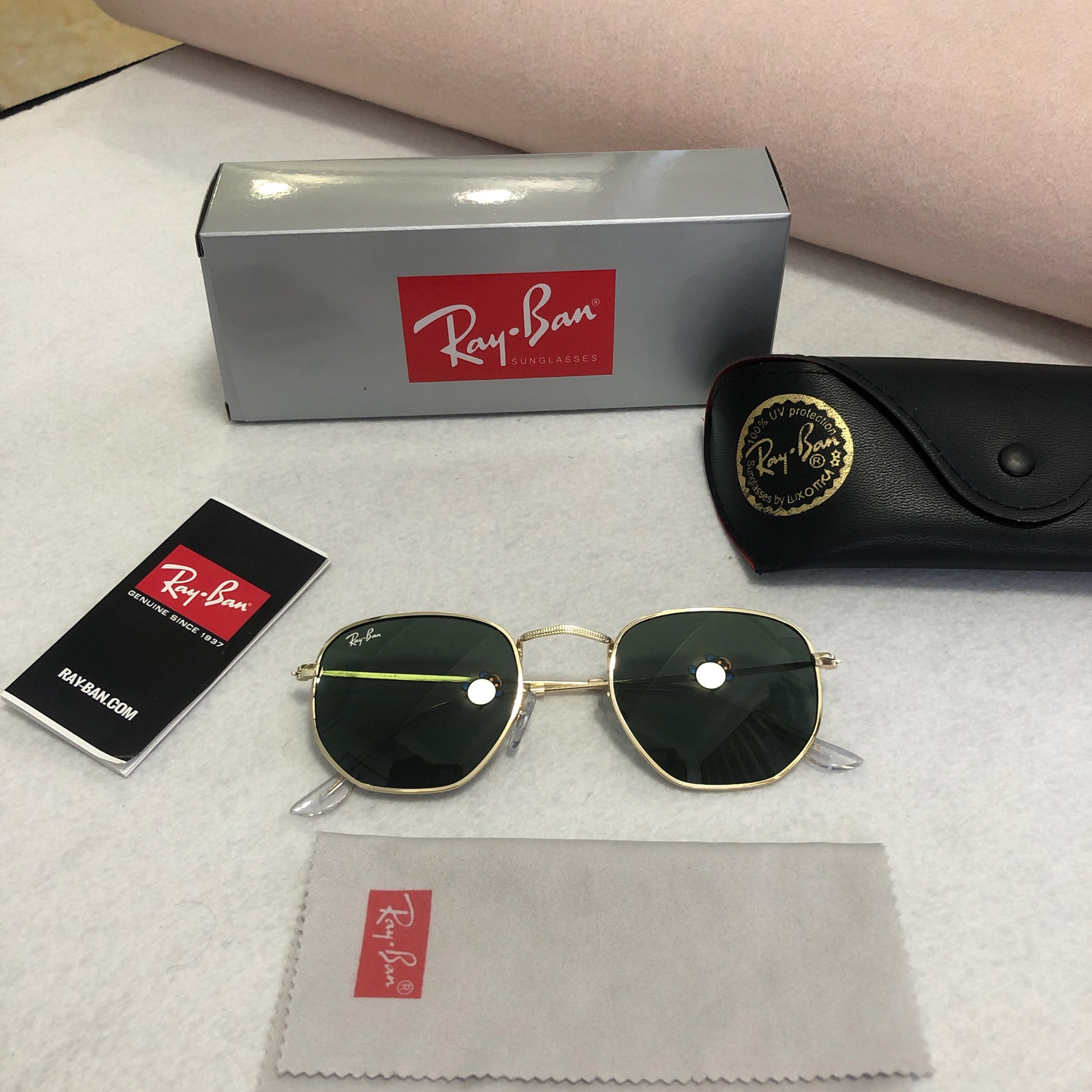 Ray-Ban sunglasses with dark green lenses and gold metal frames, displayed with their original gray box, black case, black card, and gray microfiber cloth on a light gray surface.